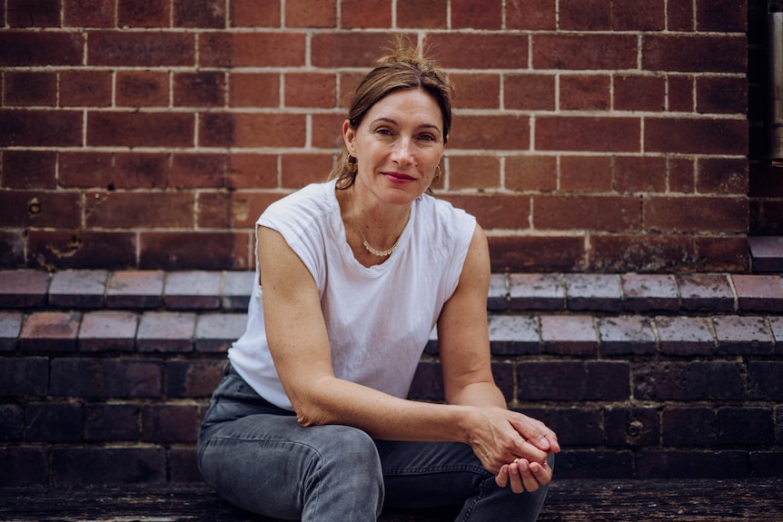 Woman in a white T shirt and jeans gives a slight smile whilst sitting in front of a red brick wall