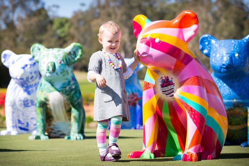 A little girl walks next to a rainbow-coloured koala sculpture.