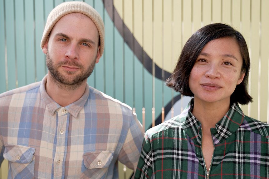 A young man and an Asian Australian woman with a slight smile, each wearing checked shirts, look into the camera 