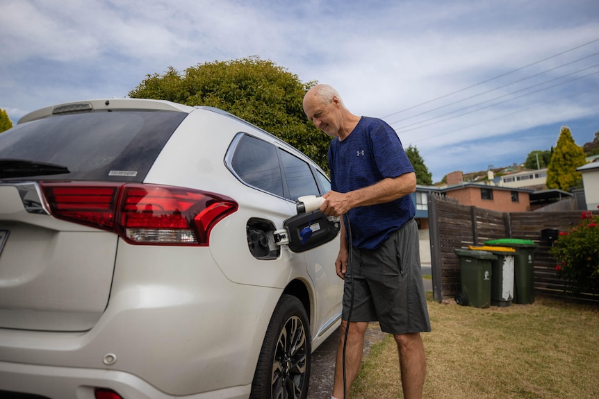 a middle aged man plugs in his white hybrid vehicle in a suburban driveway