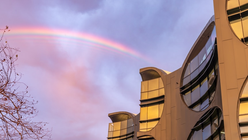 You look up at a wave-like building set against a pink/blue sky with a rainbow appering on the horizon.