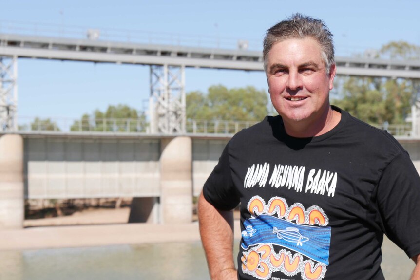 A man wearing a black t-shirt smiles, standing in front of weir gates.