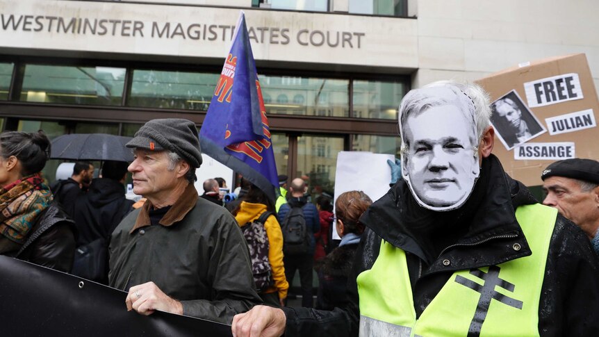 A group of supporters outside the courtroom hold flags. One man wears an Assange mask.