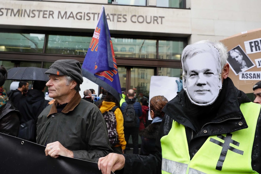 A group of supporters outside the courtroom hold flags. One man wears an Assange mask.