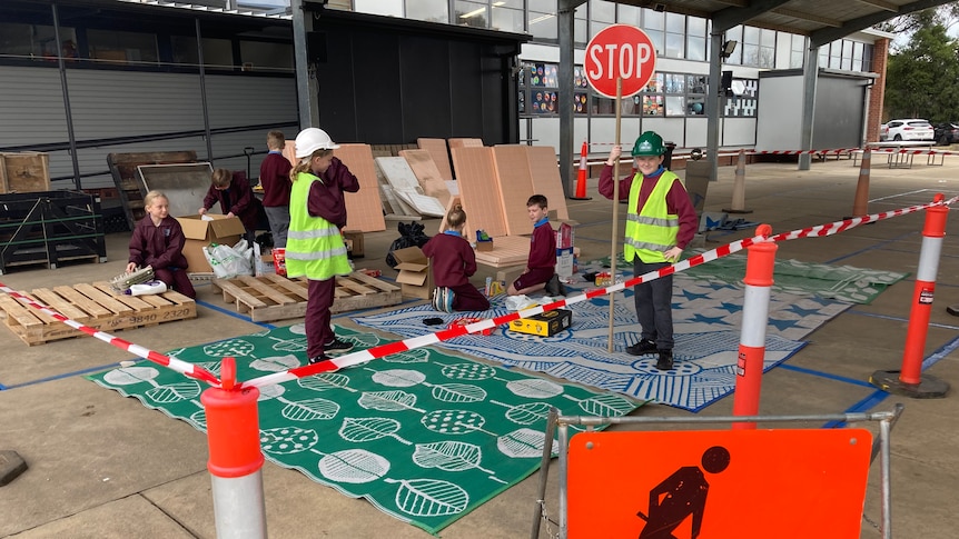 Students stand around a mock 'construction zone', some wearing hardhats and fluorescent vests, surrounded by cardboard boxes.