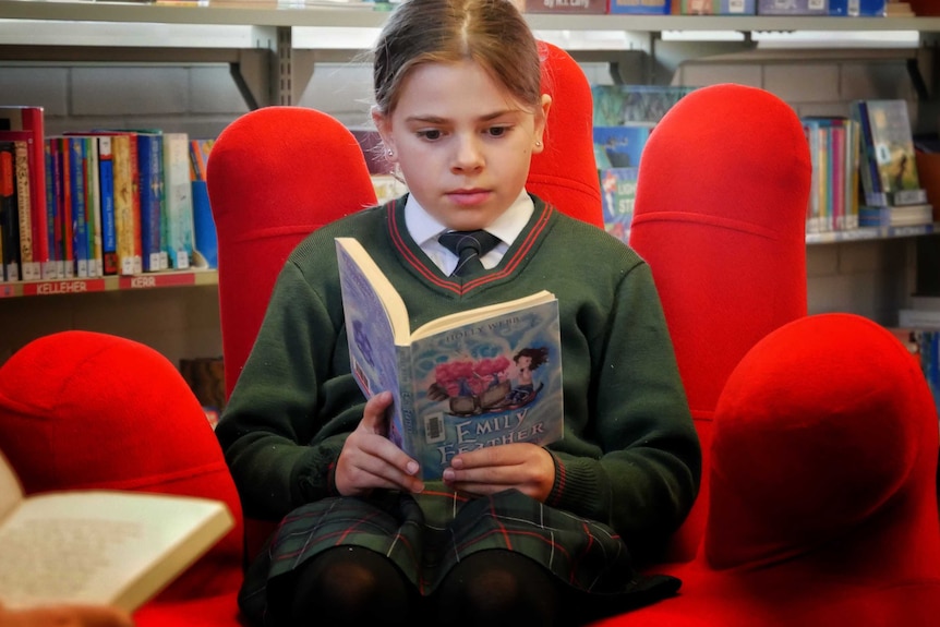 A female student from Mel Maria Catholic Primary School sits in a giant red chair in a library reading a book.