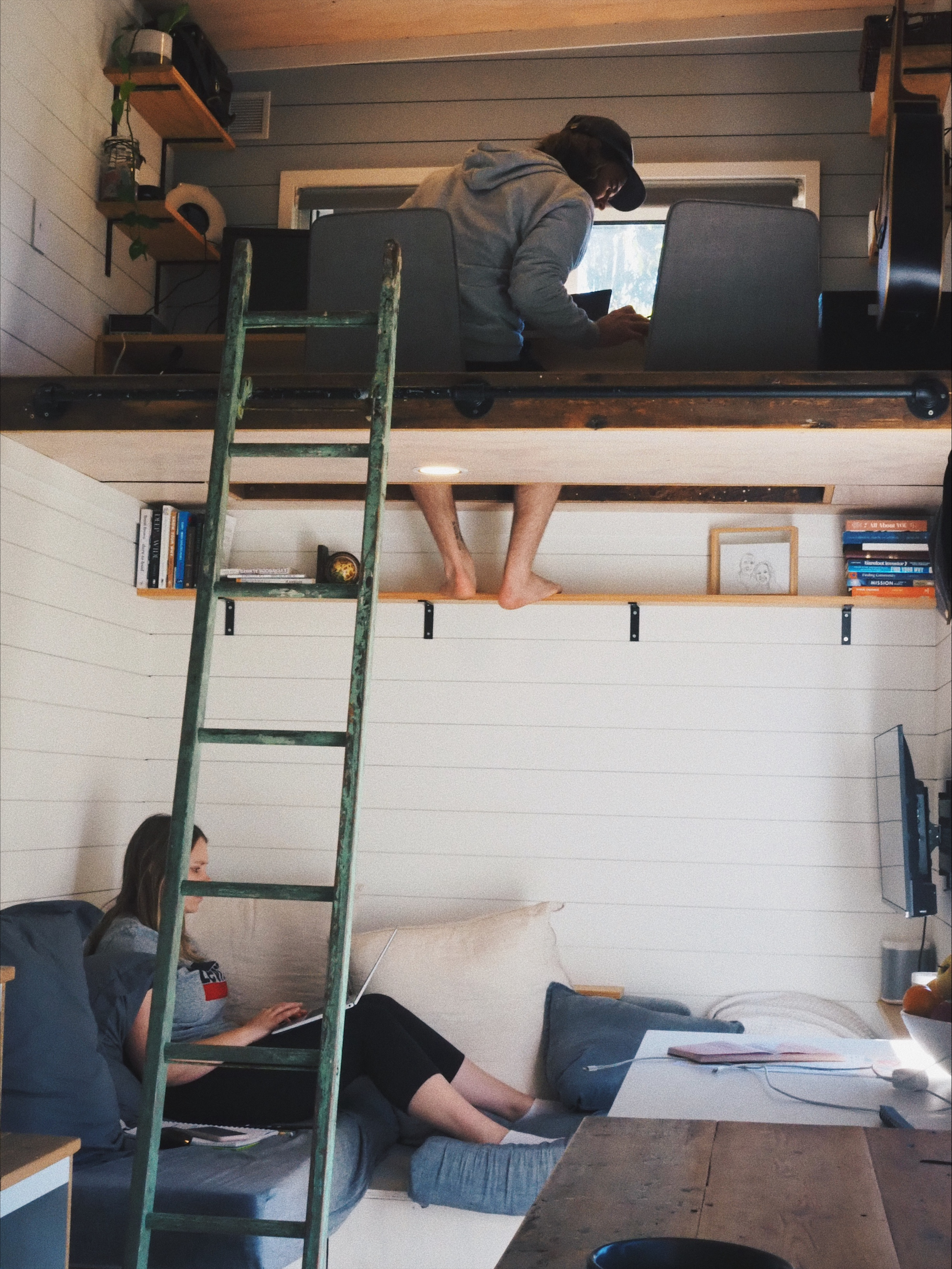 A young woman sitting on a couch works on her laptop while above her on a wooden platform a young man sits in a makeshift study.