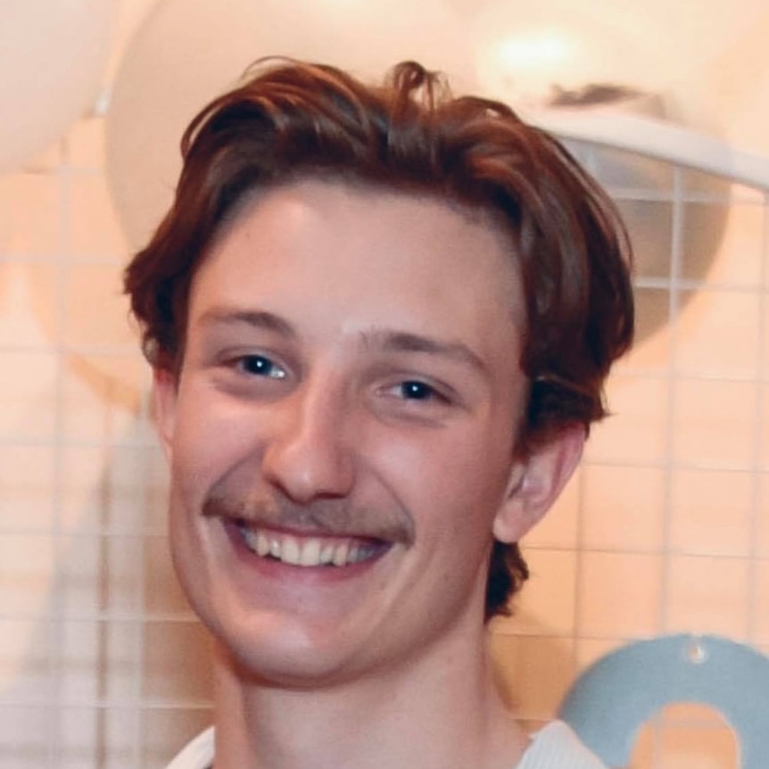 A young, brown-haired man with a mustache smiles at the camera 