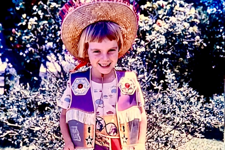 A young Louise smiles at the camera, wearing colourful cowgirl outfit and hat.