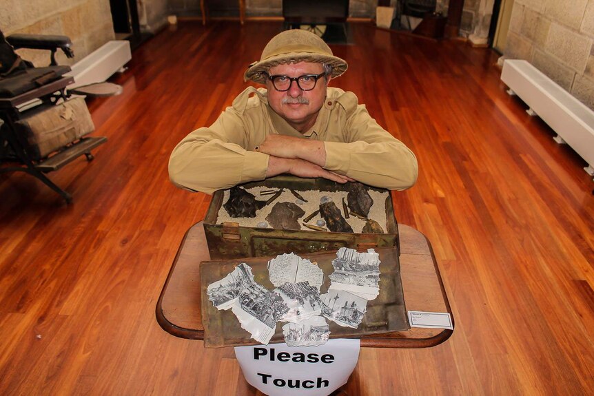 A man in a replica tin army hat rests his hands on an army supplies box filled with sand and bullets