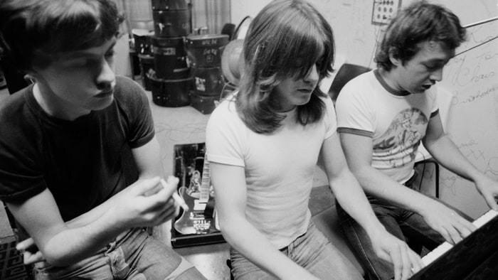 AC/DC producer George Young sitting at a piano with brothers Malcolm and Angus Young.