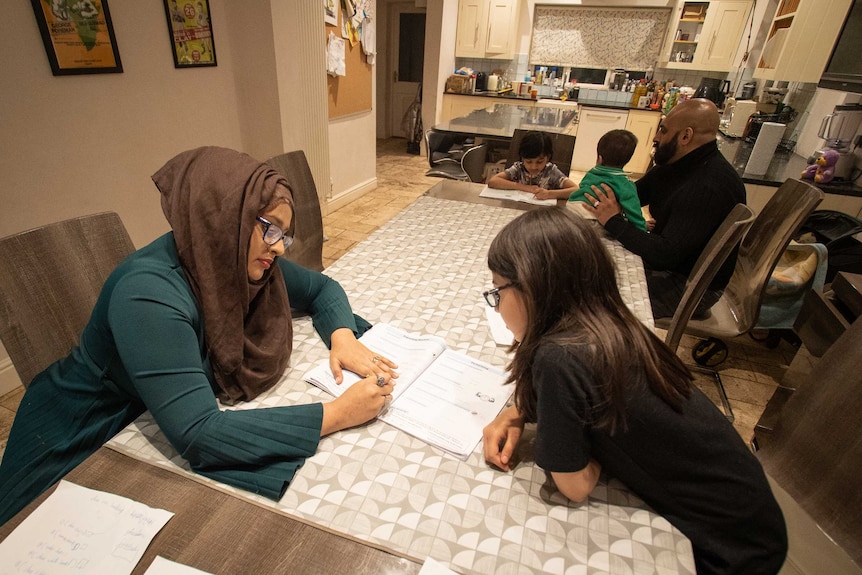 A mother works with her daughter at the dining table while a father looks after two other children.