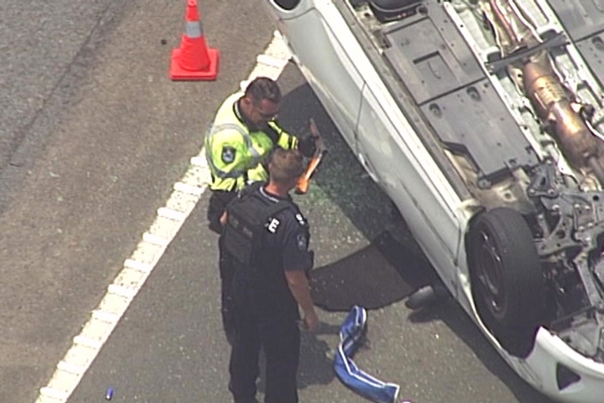 An aerial photo of a police officer holding what appears to be a shotgun in front of an upturned white sedan.