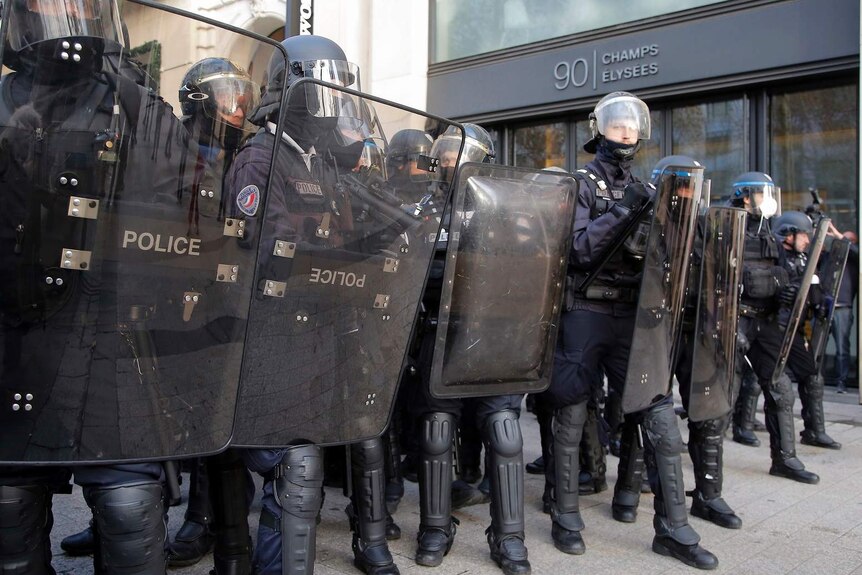 Police in riot gear hold shields to form a lint on a street marked 90 Champs Elysees