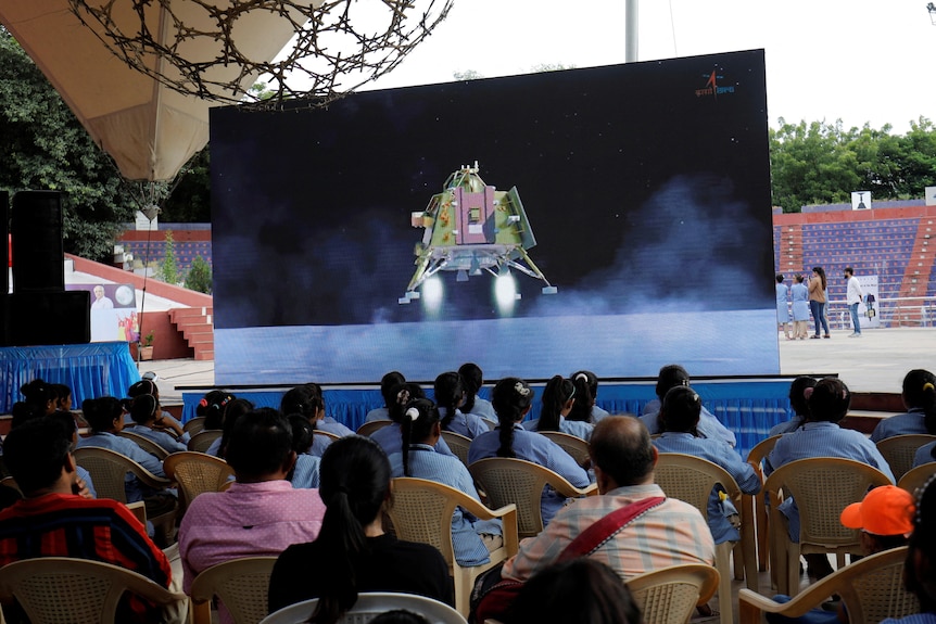 People seated watch screen broadcasting India moon mission.