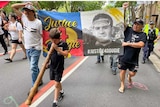 Protesters carry a large banner saying 'justice for Dougie' while a young boy walks in front playing a didgeridoo 
