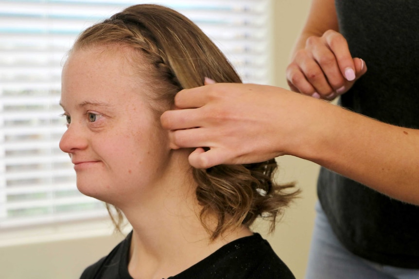 A woman plaits Sarah's hair as she looks into a mirror.