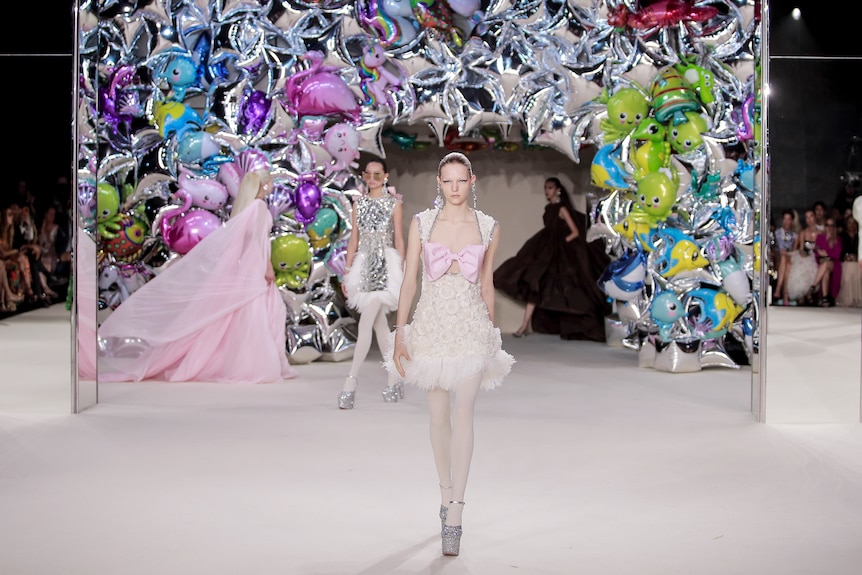 A model in a white dress with a pink bow in front of a balloon arch with three models in the background.