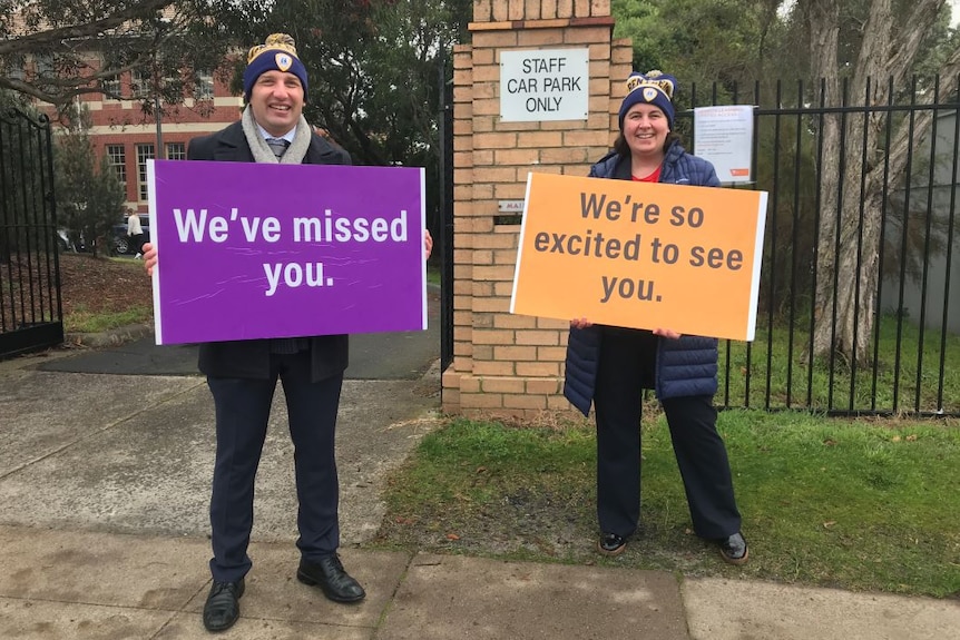 A man and a woman stand outside a school holding signs saying 'We've missed you' and 'We're so excited to see you'.