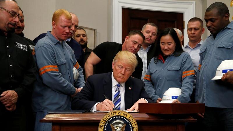 Donald Trump signs the document at his desk surrounded by workers.