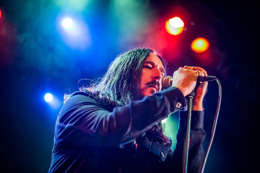 A man with straight, long dark hair holds a microphone under bright lights.