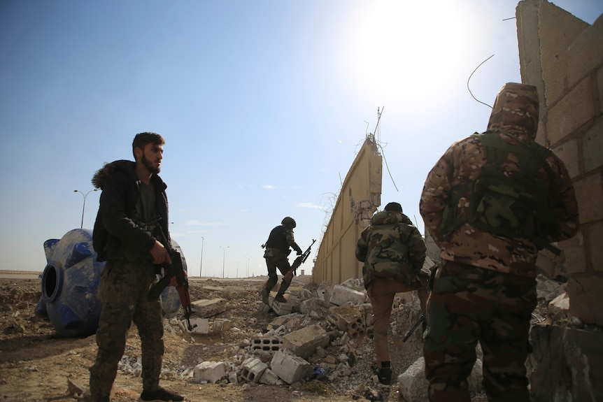 Men with big guns in camo gear stand around a hole in a fence 