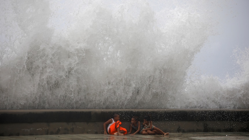 three small children hide behind a low sea wall as a massive wave breaks behind them