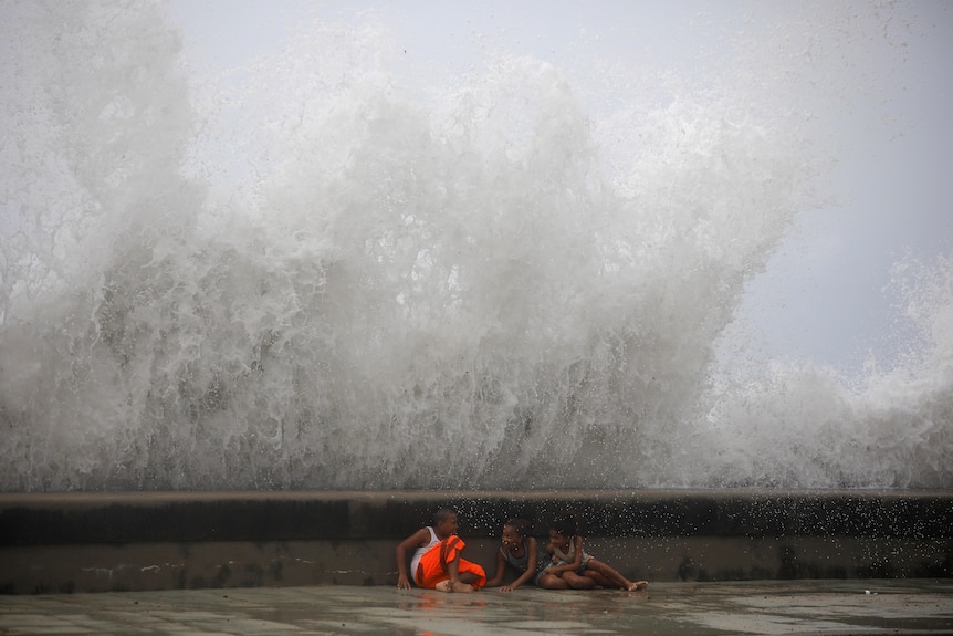 three small children hide behind a low sea wall as a massive wave breaks behind them