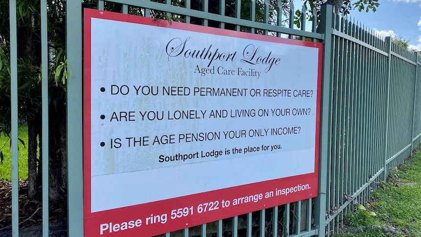 A sign about Southport Lodge Aged Care Facility pinned up on a green fence with green grass below and blue sky above.
