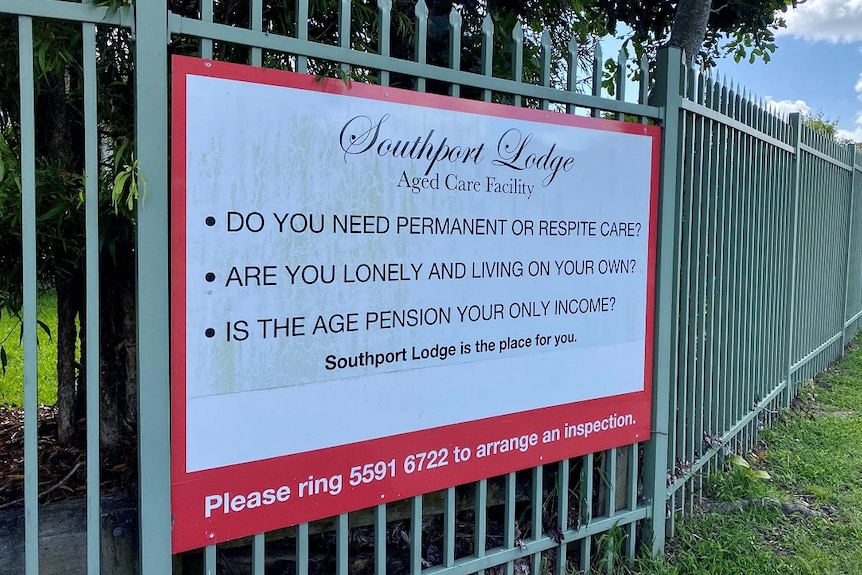 A sign about Southport Lodge Aged Care Facility pinned up on a green fence with green grass below and blue sky above.