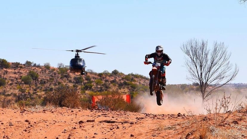 A motorcyclist soars through the air over a sand dune, with a helicopter behind him.