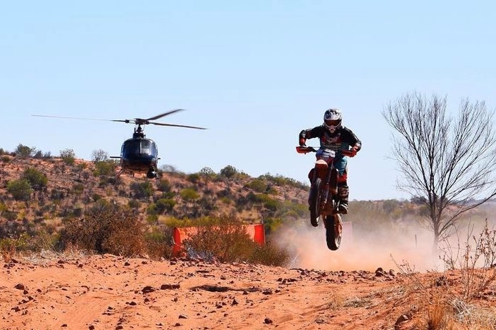 A motorcyclist soars through the air over a sand dune, with a helicopter behind him.