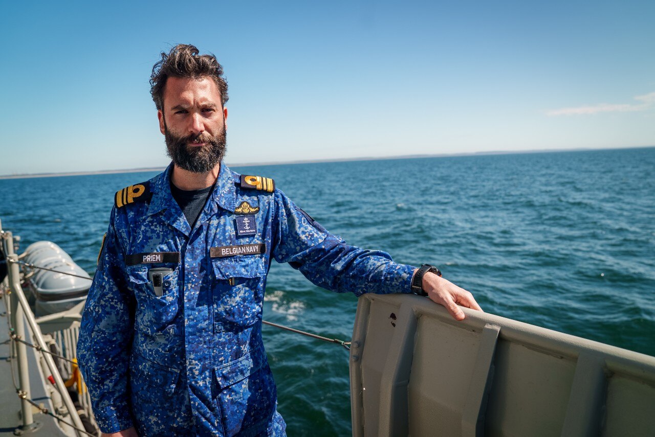 A man in a naval uniform, pictured on the deck of a ship, looking on.