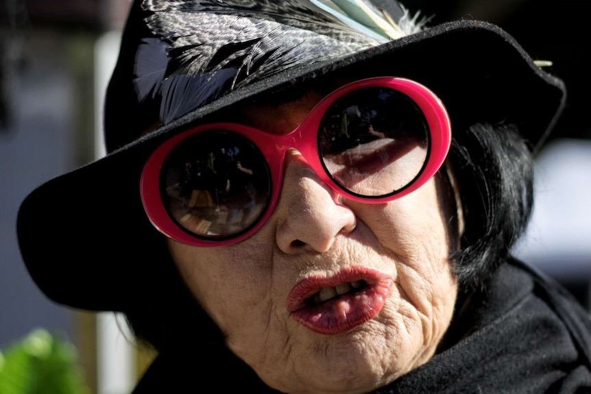 A woman in a black-hat, red lipstick and red-rimmed sunglasses is photographed mid-speech.