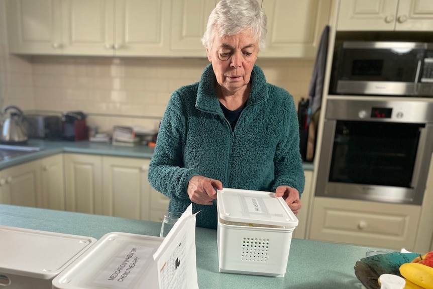 Lyn Read wearing an aqua green sweater, is standing behind a kitchen counter, with boxes of her medication atop the bench.