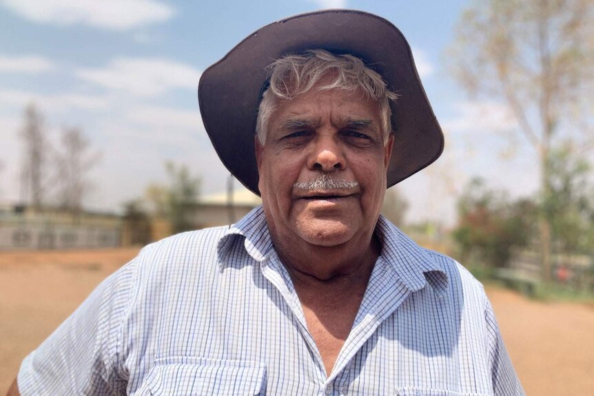 An older, moustachioed man in a hat stands in a desert landscape.