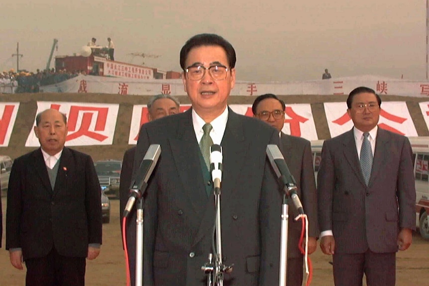 An old photo shows Li Peng behind three microphones on a red-carpet ceremony with a row of five male officials behind him.