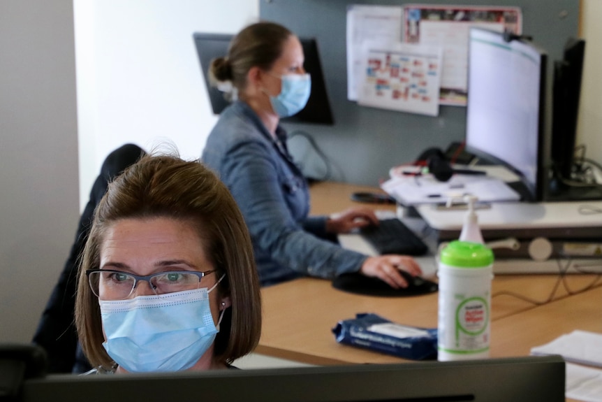 A woman with short hair wearing a mask and glasses stares ahead , concentrating. Another woman can be seen at a computer behind.