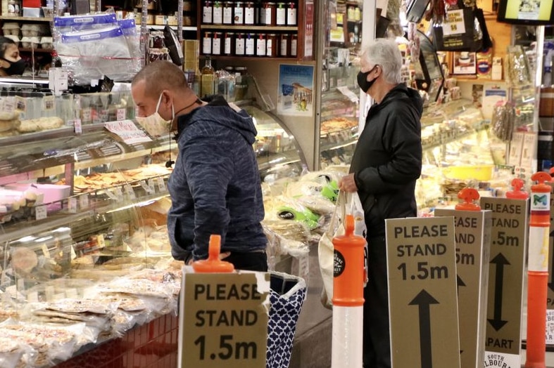 Two shoppers in masks wait at the counter of a deli stall in an indoor market.