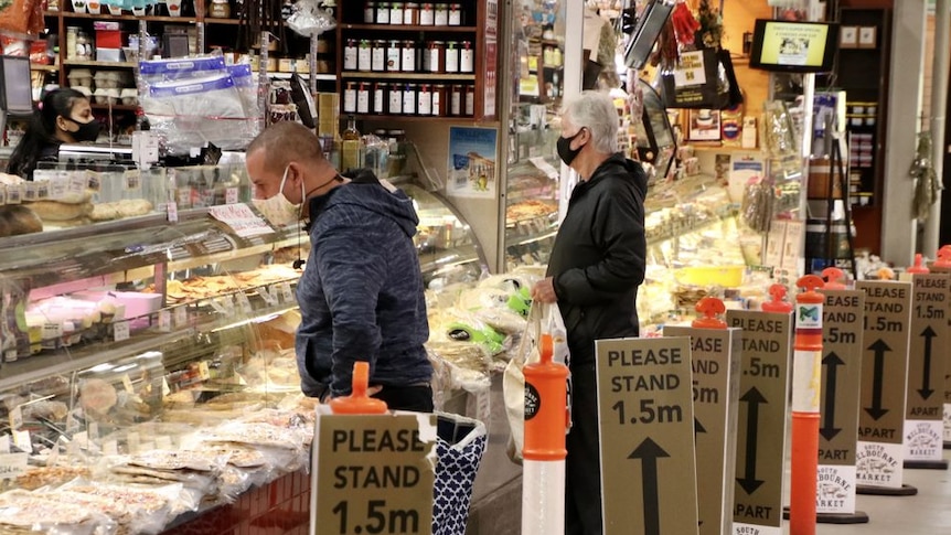 Two shoppers in masks wait at the counter of a deli stall in an indoor market.