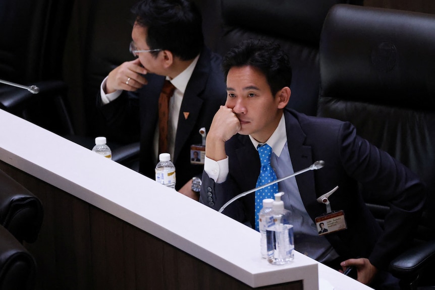 A young Thai man in a suit sits at a desk, he looks glum as he rests his head on his hand