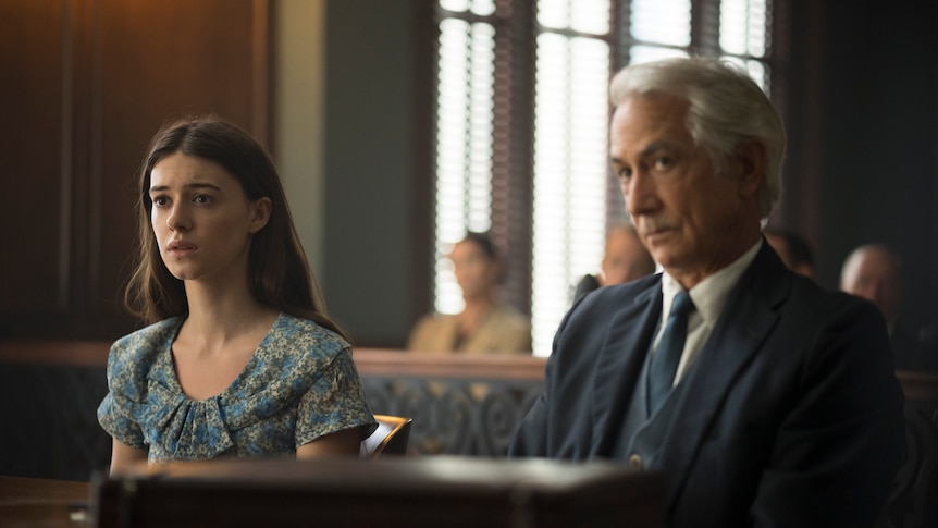 A long-haired brunette sits with a distressed expression behind a desk in a courtroom, beside an older man in a suit