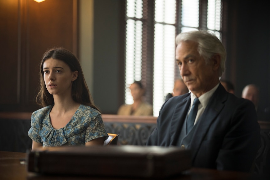 A long-haired brunette sits with a distressed expression behind a desk in a courtroom, beside an older man in a suit