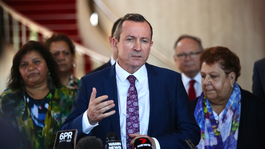WA Premier Mark McGowan dressed in a suit speaks at a lectern watched on by other people. 