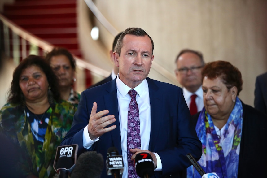 WA Premier Mark McGowan dressed in a suit speaks at a lectern watched on by other people. 