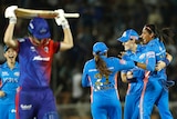 Meg Lanning, blurry in the foreground, walks off with her bat over her head as Mumbai Indians players celebrate her wicket.