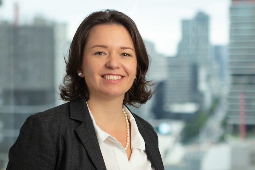 A smiling, formally dressed woman with dark hair standing in a high-rise office building overlooking a city.