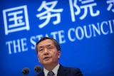 A Chinese man in front of a blue screen with chinese writing on it and "the state council" 