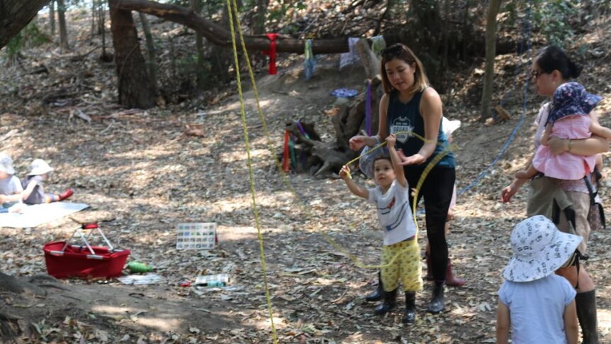 Parents standing around and playing with  children using ropes in a bush clearing