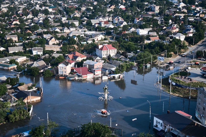 An aerial shot shows flooded streets leading to a flooded open area with a statue on a column.